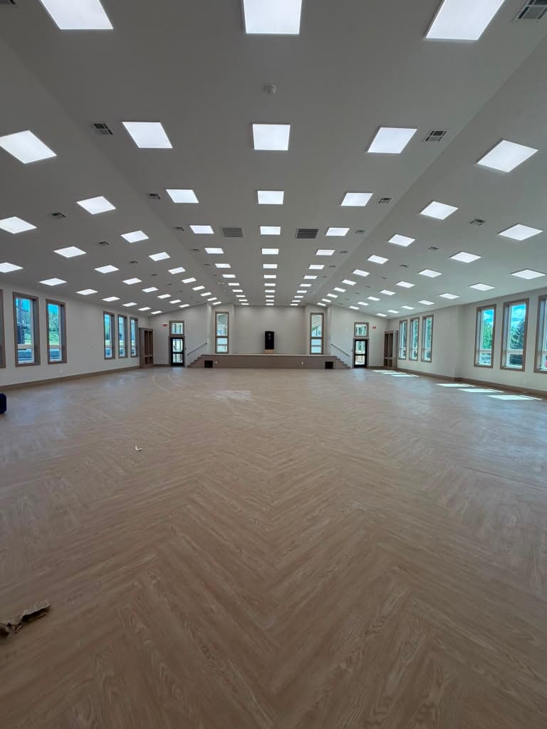 A wide clear-span assembly hall, empty, with a raised stage and steps at the far end and herringbone flooring run the length of the room