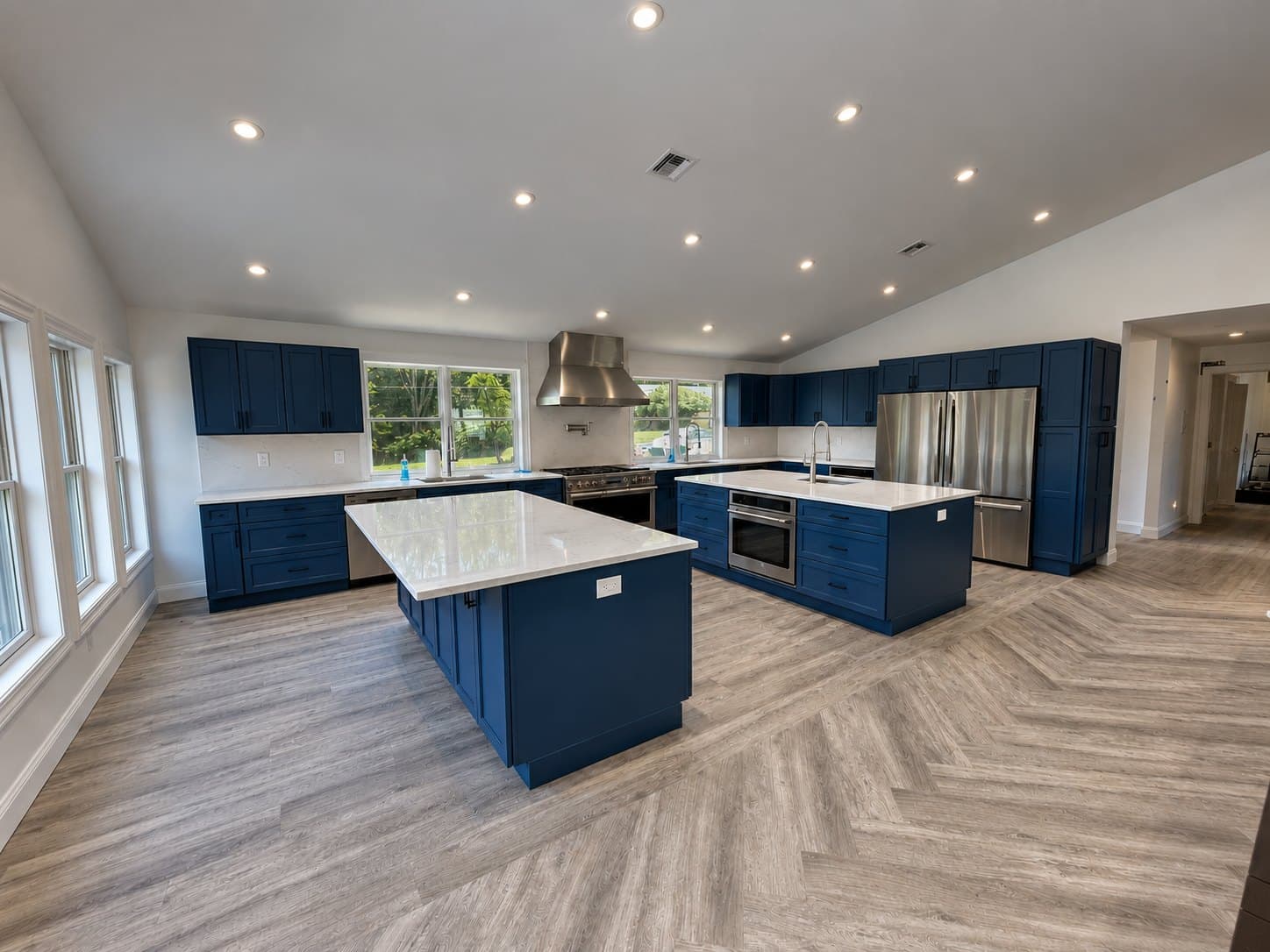 A kitchen with navy cabinetry, a long island with a stone top and herringbone flooring under a vaulted ceiling
