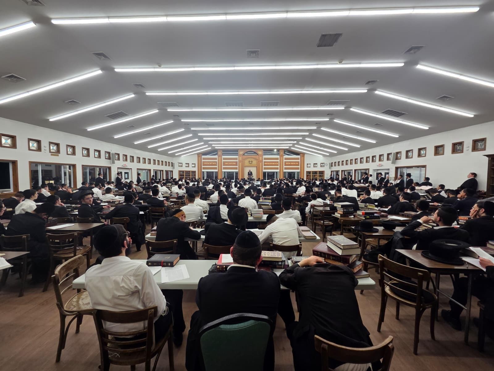 A beis medrash in use, rows of study tables running the length of the room with sefarim open on every table, under continuous ceiling light set in a chevron