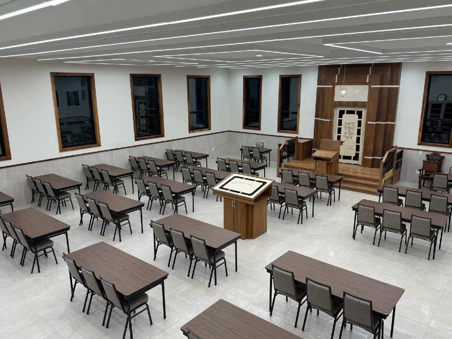 The interior of a new beis medrash, tables and chairs set around a central bimah with the aron kodesh and a raised platform along the far wall