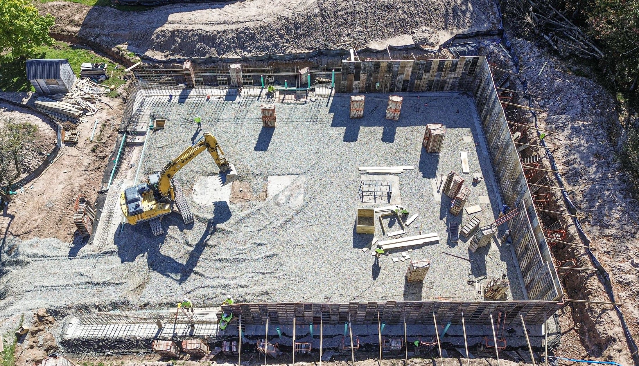 Drone view straight down into a shul's foundation, poured walls framing a level gravel pad with an excavator working inside