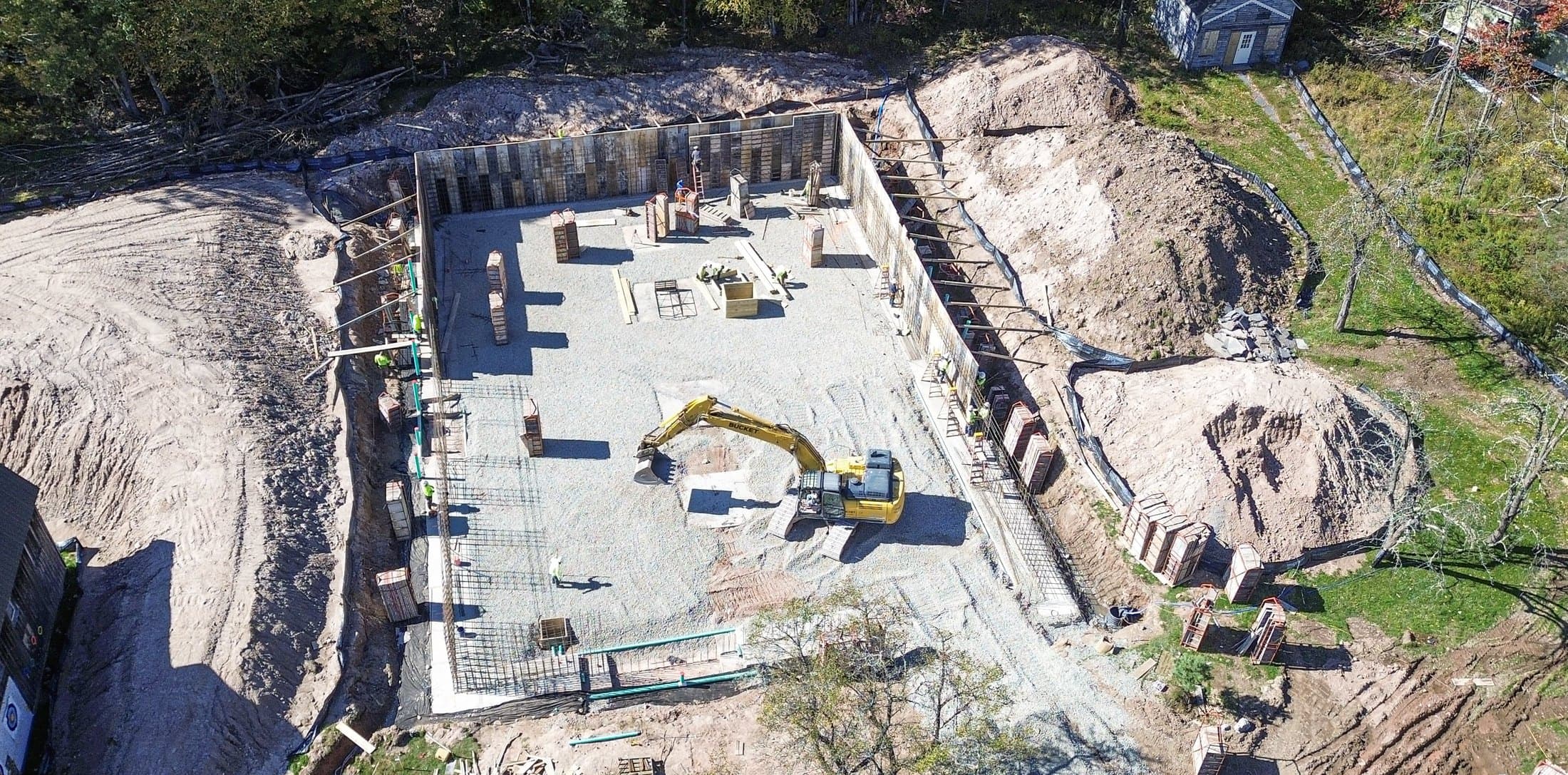 Wide aerial of a finished foundation and its stone pad, with the excavation cut back into the trees around it