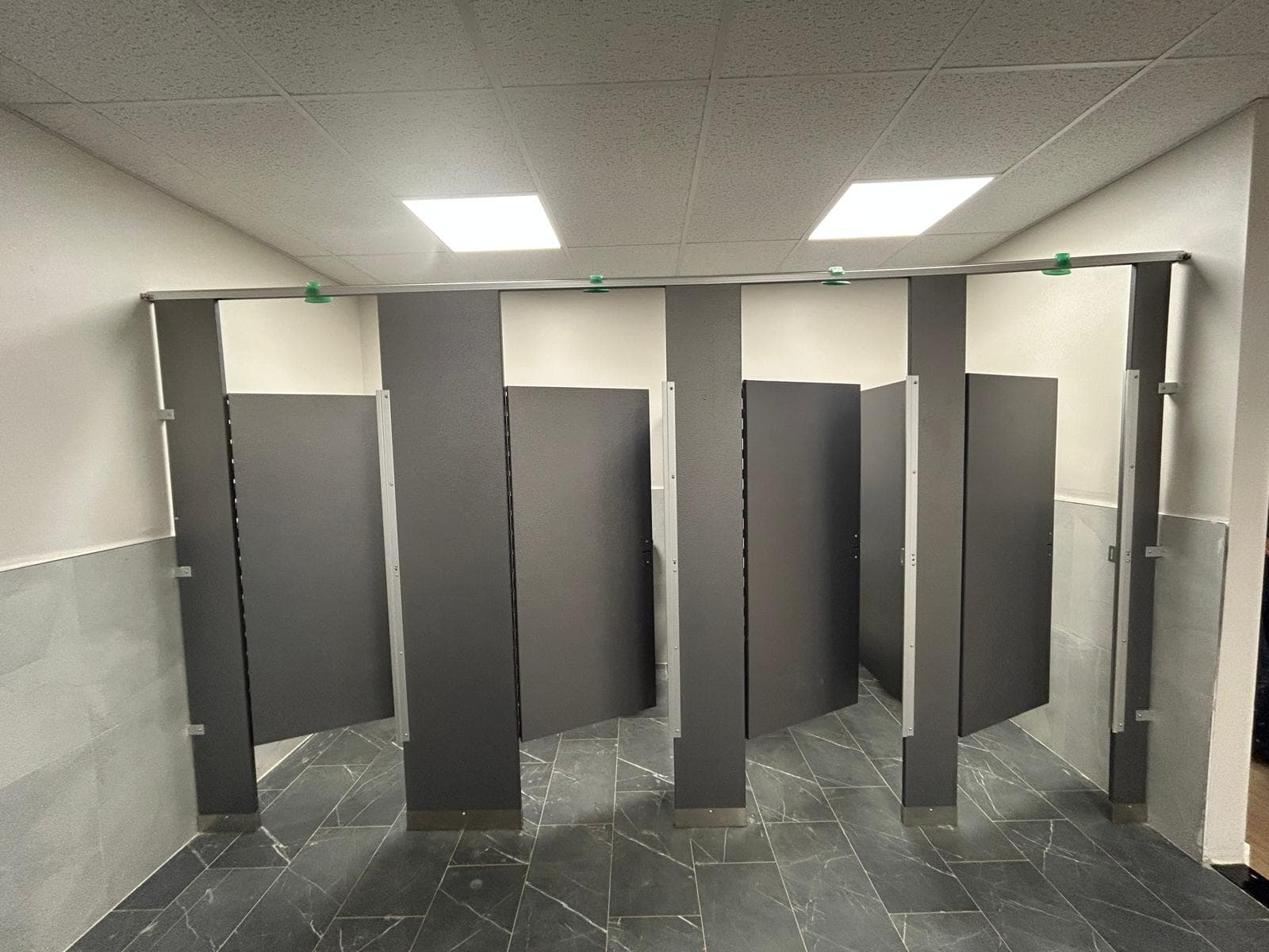 A row of floor-mounted washroom cubicles in dark laminate, tiled half-height walls behind them